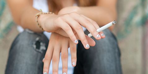 Hände einer weiblichen Jugendlichen mit lackierten Fingernägeln. Sie hält eine Zigaretteich der rechten Hand. Die Hände sind übereinander geschlagen. 