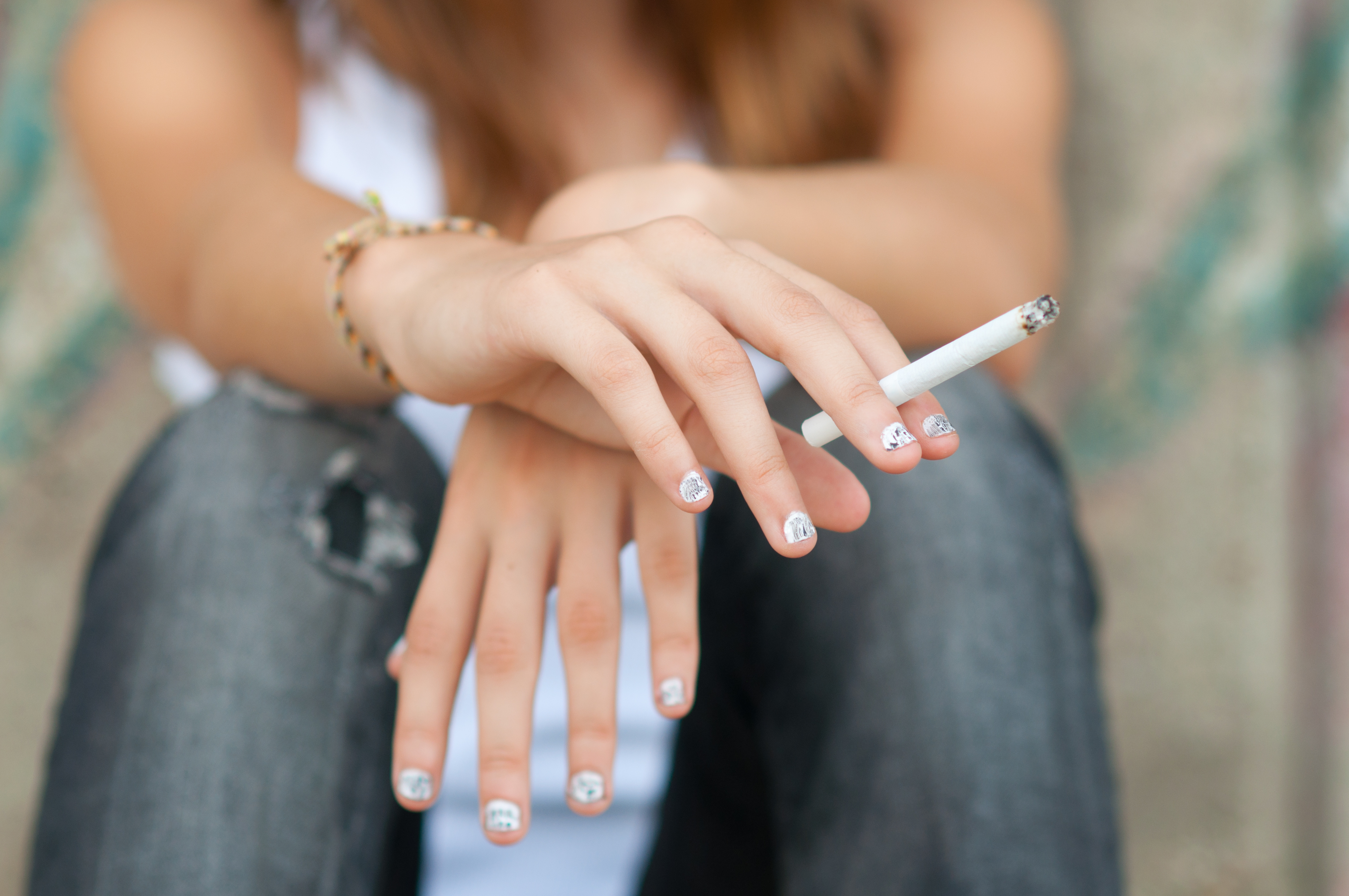 Hände einer weiblichen Jugendlichen mit lackierten Fingernägeln. Sie hält eine Zigaretteich der rechten Hand. Die Hände sind übereinander geschlagen. 